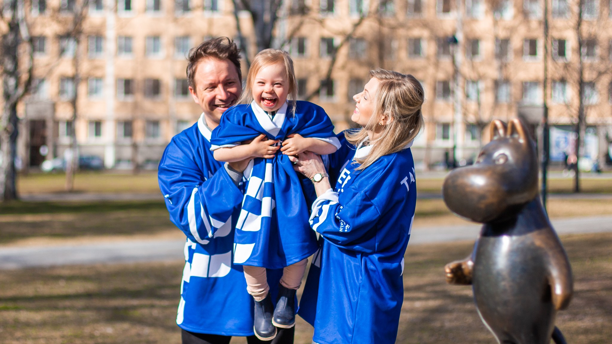 Äiti, isä ja lapsi muumipatsaan kanssa puistossa iloisina MM-jääkiekkopaidat päällä.