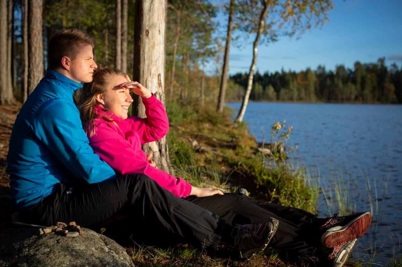 Nuoripari istuu sylikkäin järven rannalla ja katsoo horisonttiin.