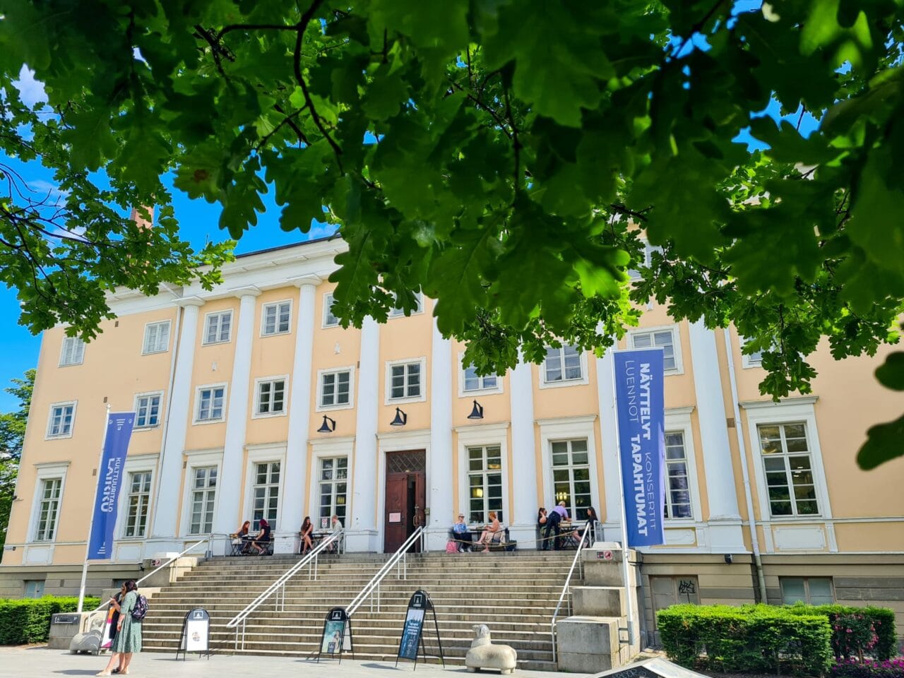 Iso portaikko johtaa keltaiseen rakennukseen. Portaikon ylätasanteella istuu ihmisiä kahvipöytien ääressä.