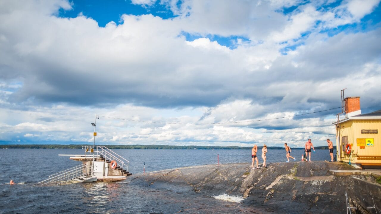 Järvimaisema, sinisellä taivaalla pilviä, uimareita menossa laituria kohden uimaan