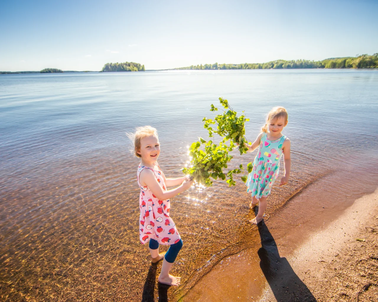 Kaksi lasta rantavedessä, joilla on saunavihdat kädessä.