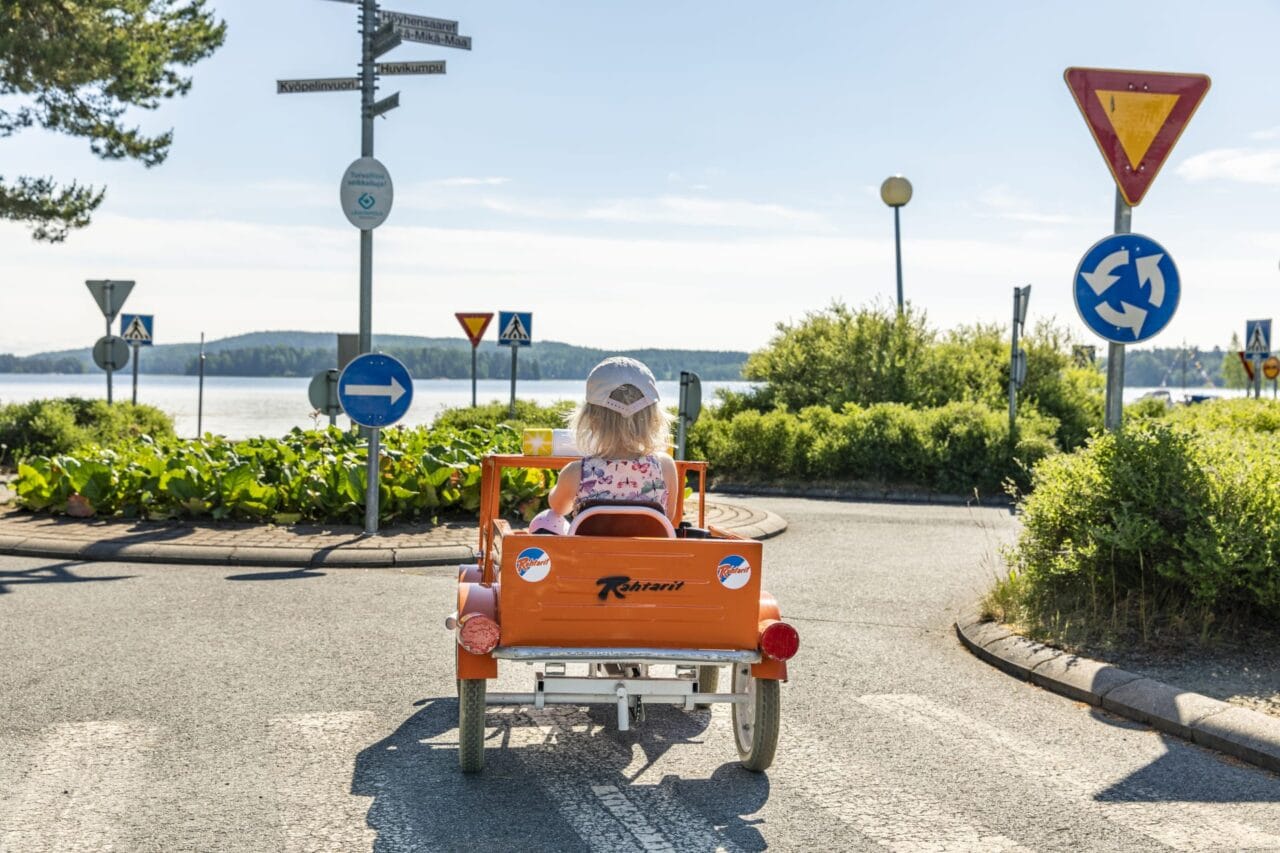 Lippalakkipäinen tyttö ajaa oranssilla polkuautolla liikenneympyrään. Taustalla järvi.