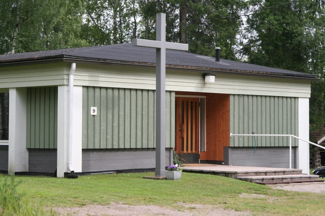 Kesällä otettu valokuva Länsi-Teiskon rukoushuoneesta. Kohde on melko iäkkään näköinen, matala vaaleanvihreäksi maalattu puurakennus, jonka edustalla kohoaa katon korkeuteen ulottuva risti.