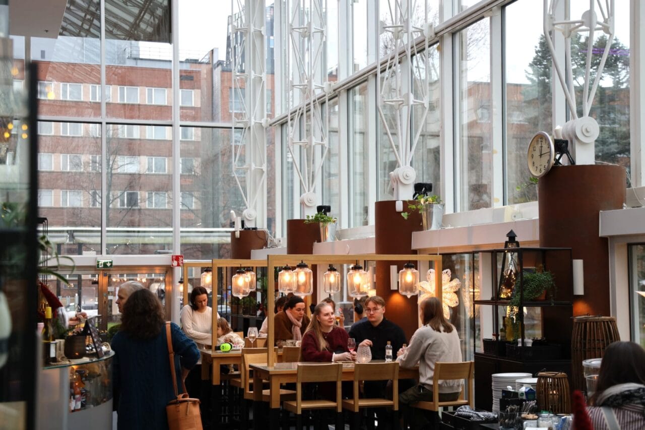 Valoisa ravintolatila, jossa asiakkaat ruokailevat suurten ikkunoiden äärellä. / Bright restaurant interior with guests dining by large windows.