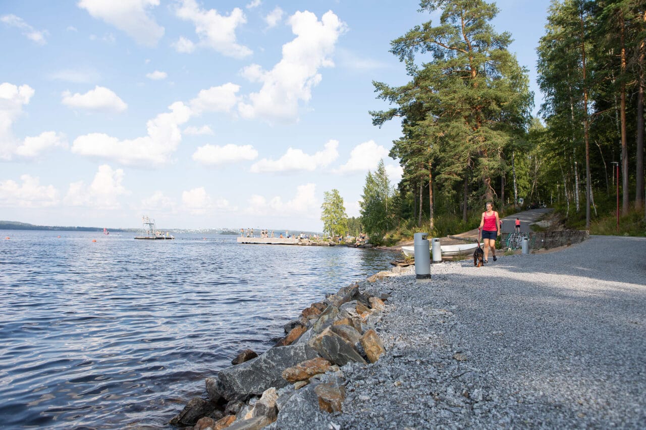 Henkilö kävelee koiran kanssa rannalla. Taustalla uimaranta.