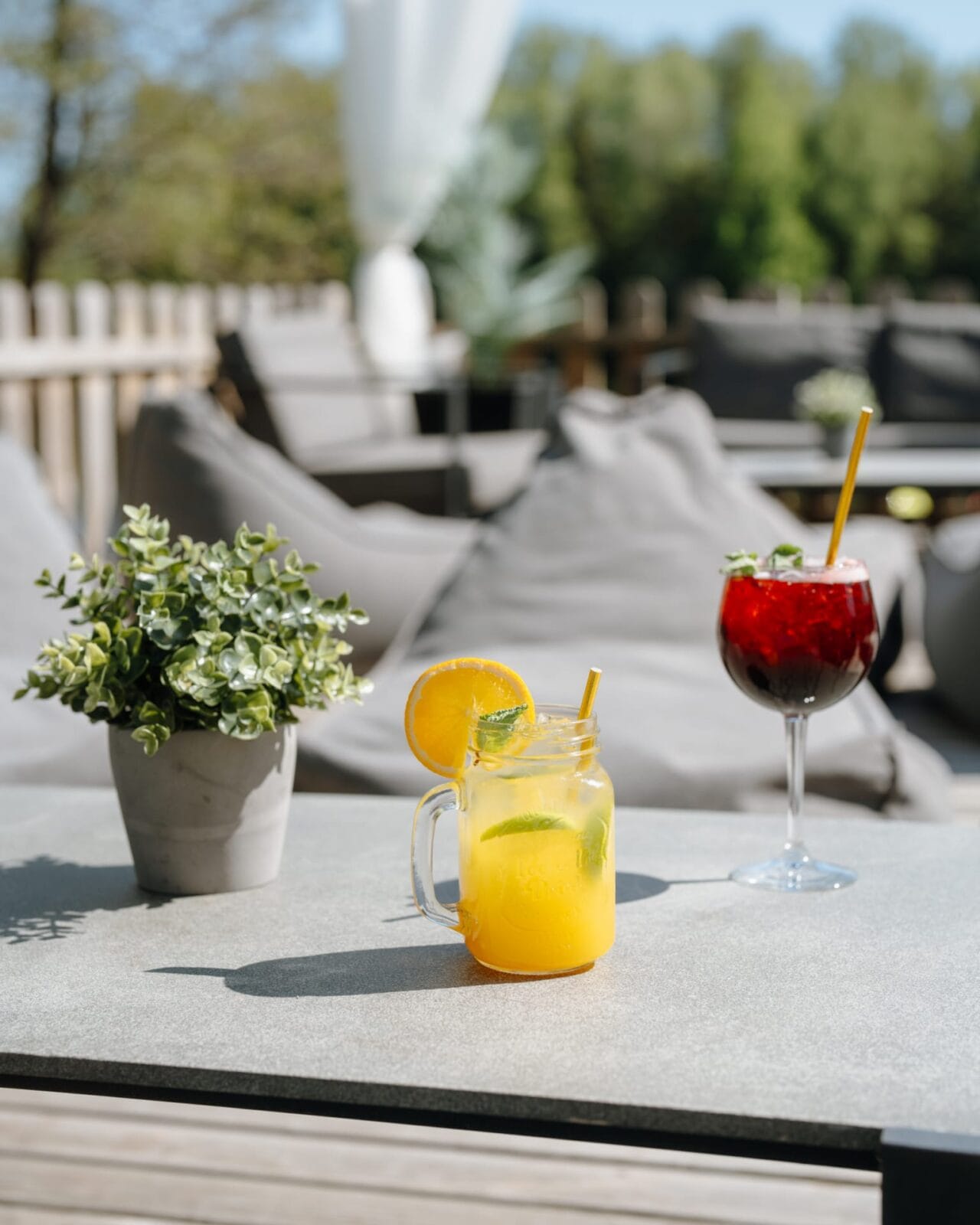 Two refreshing cocktails on an outdoor table: a yellow drink in a mason jar garnished with an orange slice and mint, and a red drink in a wine glass with a straw, set against a background of outdoor lounge furniture and greenery