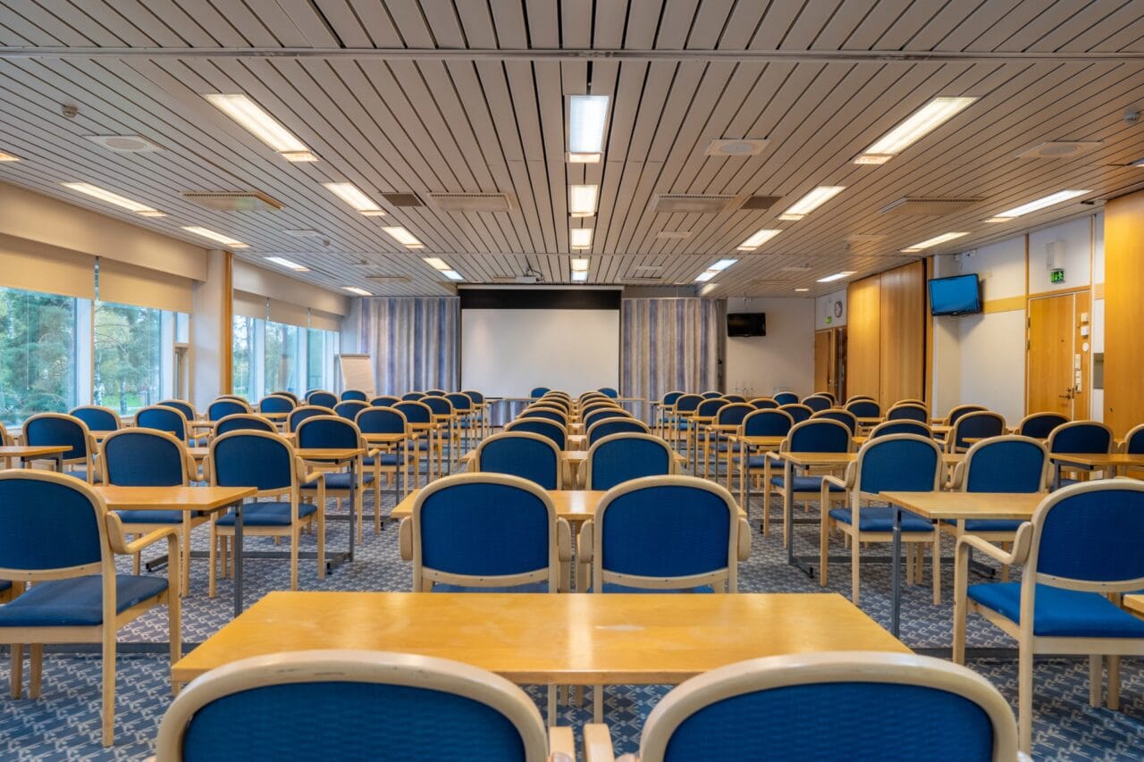 A large conference room set up for a presentation, featuring rows of light wood desks and matching armchairs with blue upholstery facing a projection screen at the front of the room.