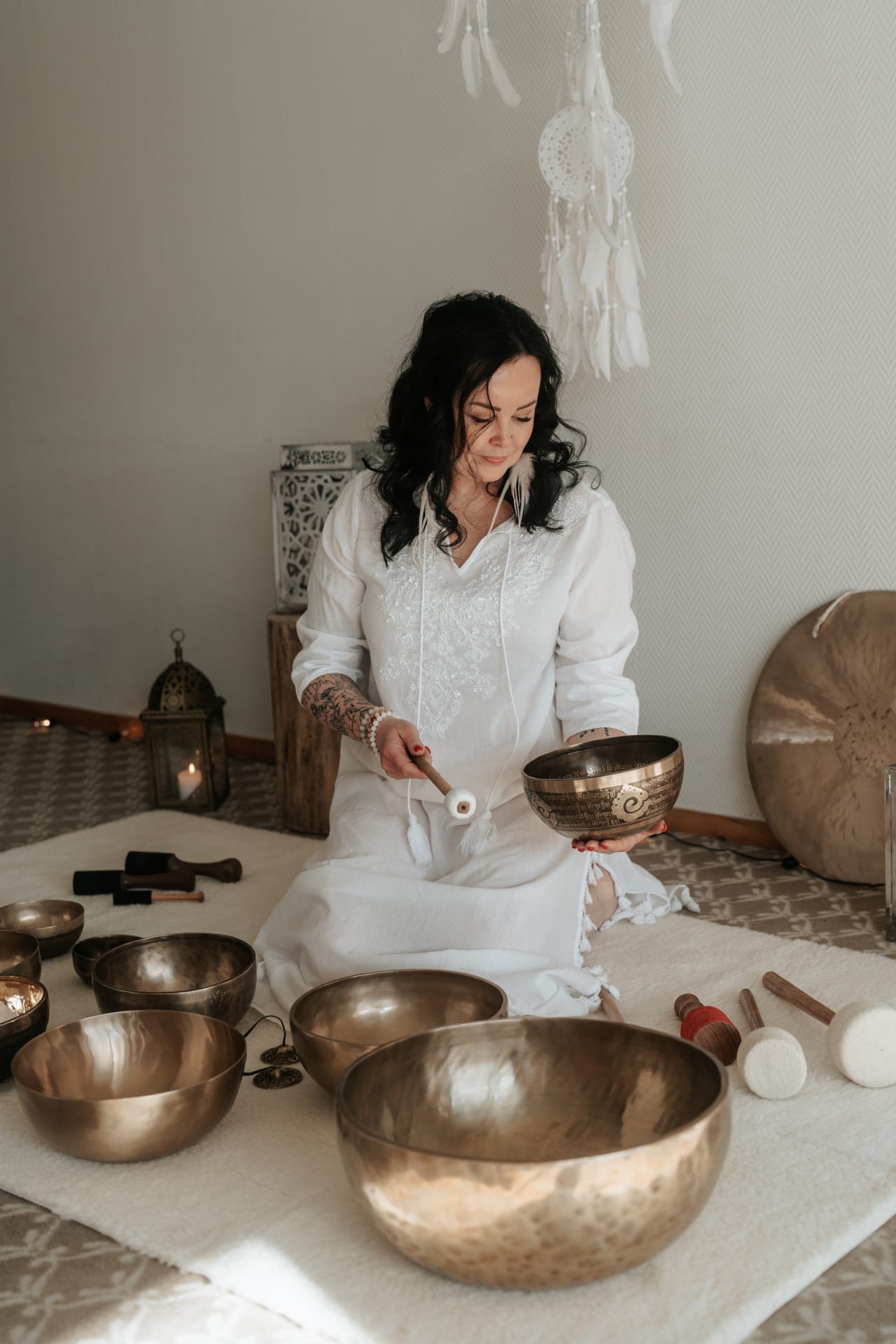 A woman in a white tunic sitting on a white rug, holding a singing bowl and a mallet, surrounded by several other Tibetan singing bowls, mallets, a lantern, and a dreamcatcher hanging in the background.